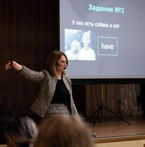 Woman in gray suit standing in front of an audience teaching English lesson