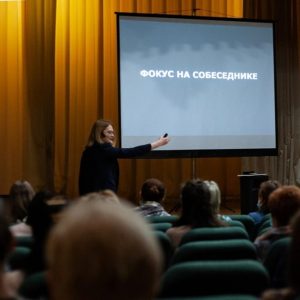 Woman in black suit standing in front of audience pointing to presentation